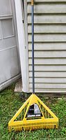Photo shows a large yellow yard rake with a triangular head and multiple yellow tines attached to a long handle leaning against siding.