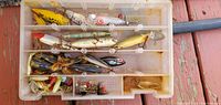 Top view of plastic tackle box with assorted fishing lures, showing 17 used lures of various types and colors in individual compartments