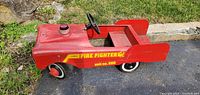 Side angled view of the red vintage AMF pedal car showing 'FIRE FIGHTER unit no. 507' decal with visible dents on hood and rear spoiler-like back ends.