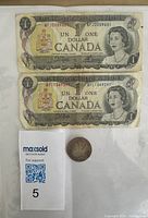 Front view of two Canadian one dollar bills from 1973 Series BF with Queen Elizabeth II, placed above a German coin and a MaxSold identifier card.
