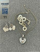 Silver bracelet with circular links and toggle clasp, pair of earrings with coin charms, and heart pendant necklace laid out on a gray surface.