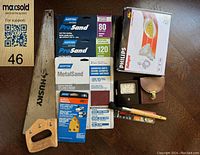 Photo of various hand tools and sanding papers including Husky hand saw, Norton sanding papers, Philips lamp bulb, analog meter, and drill bit.
