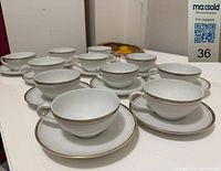View of 11 white porcelain tea cups and saucers arranged on a table, showing the set and gold detailing.