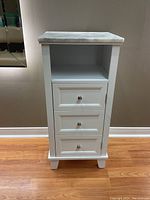 Front view of the white cabinet showing three drawers with silver knobs, open shelf, and marble top.