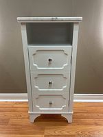 Front view of the white cabinet with marble top, three decorative drawer-style panels on the door, each panel has a chrome knob, showing cabinet in closed position with visible wood grain flooring and wall background.