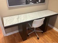 Desk with a marble top and dark legs, shown against a wall with a framed picture above it. Chair with white molded seat and silver base on casters placed under desk.