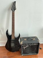 Black Washburn electric guitar standing upright next to a Fender Frontman 10G amplifier on wooden floor against white wall.