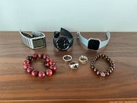 Display of all items on a wooden surface, showing three watches, four rings, and two bracelets.