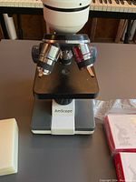 Close-up of AmScope microscope showing rotating three objective lenses and stage with clips; instrument stands on table with keyboard visible in background