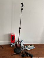 Image showing the full lot against a white wall on a wood floor. Microphone stand in center, unopened Bluetooth Music Receiver box, headphones, game controller, power strip, and cables arranged around it.