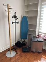Wide angle showing all items together: two coat racks, ironing board, two trash cans, body scale, and clothes hangers on hardwood floor near shelves and window with blinds.