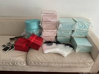 Image showing assortment of decorative gift boxes and packaging supplies arranged on a beige couch, featuring blue, pink, and red boxes, chalkboard tags, and clear sleeves.
