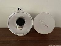 Front view of Samsung Gear S2 smartwatch in its round white box with lid beside it, showing the watch face and band.