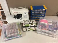 Wide shot: Singer One sewing machine with power adapter, blue/yellow multi-drawer organizer, clear plastic bead organizer, two clear plastic craft storage boxes and fabric-covered sewing kit box