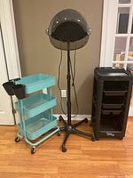 Black hooded hair dryer on adjustable stand, turquoise cart with shelves and black cylindrical holders, and black rolling storage cart all positioned side by side on wood floor.