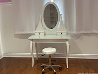 Front view of white vanity table with glass top, oval mirror, two small drawers and rolling stool in front.