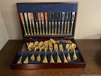 Full view of the Wallace gold-tone cutlery set in the wooden presentation box showing knives arranged in the lid and forks, spoons, and serving pieces in the bottom section.