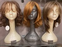 Three wigs on mannequin heads: light brown layered with bangs, bright copper layered, and light brown with waves