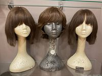 Three mid-length brown synthetic wigs displayed on two cream-colored and one silver styrofoam mannequin heads.