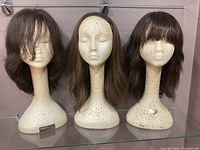 Overview showing three wigs on off-white mannequin heads with textured finishes, all brown shades, two with bangs and one without.