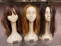Three Styrofoam mannequin heads side by side, each with a different wig: medium brown wavy, long orange straight, and long black straight.