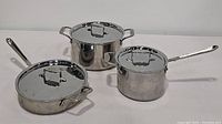 Three All Clad stainless steel pots together with lids on a white background. One pot has a long handle, one pot has two side handles, and one pot is wider and shorter with two side handles.