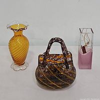 Front view of three blown glass pieces: yellow-brown ruffled vase, amber swirled handbag shape, pink abstract square vase