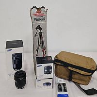 Overview showing tripod in box, two stacked Samsung lens boxes, loose telephoto lens, canvas camera bag, and remote shutter release in blister pack.