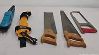 Lot items including Gardena ComfortCut cordless grass shear, Silky Sugoi saw in sheath, two hand saws with wooden handles, and Millers Falls hacksaw with red handle arranged on white surface.