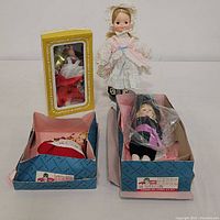 Four dolls displayed, one standing outside box in white and pink dress and bonnet, two in respective boxes, one in plastic wrap inside its box