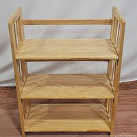 Front view showing natural wood 3-shelf folding bookshelf with simple slatted side design and three flat shelves.
