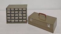 Photo showing a rectangular vintage metal tool/tackle box with a brown handle, secured by two metal clasps, and a small metal hardware storage cabinet with 30 small drawers with clear fronts and white labels.