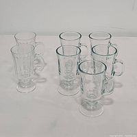 Seven clear Irish-style glass coffee mugs with handles and pedestal bases lined up on white surface.