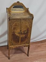 Front view of painted antique music sheet cabinet showing decorative rural scene and carved detailing.