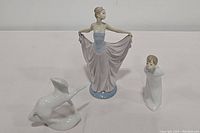 Three Lladró porcelain figurines shown including a dancer, a boy blowing, and a white animal sculpture all standing on a white background.