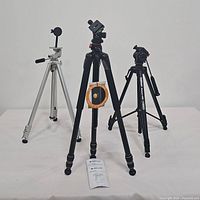 Three tripods: black Velbon Stratos 480 with video head front center, black Vanguard tripod right side, and silver metal tripod left side with mounted arm