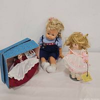 Three vintage dolls shown: one Effanbee toddler doll in blue overalls, Madame Alexander Marme #415 in pink dress standing with tag and stand, and a small doll in original box in burgundy and white dress.