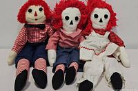 Three vintage Raggedy Ann & Andy cloth dolls displayed seated side by side showing full body and outfits.