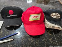 Three vintage caps on dark surface: black Island Ropes cap, red Miller High Life cap, black Jim Shockey Pro-Staff cap showing wear
