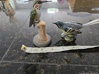 Both bird figurines side by side with measuring tape for scale, showing relative sizes, one ceramic Beswick, one hand carved wood