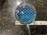 Top view of the paperweight displaying clear glass with iridescent blue bubbles inside and a tape measure for size reference indicating about 2.25 inches diameter.