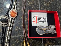 Two leather bolo ties with agate stones and a Justin Silver money clip in box on a dark granite surface.