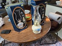 Two Lamplight Victoria Rose oil lamps on round wooden table next to original blue boxes, showing cream bases with blue flower decorations, glass chimneys, and brass burners