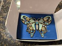 Front view of the Nina Ricci butterfly brooch made for Avon, showing light blue, aqua enamel and rhinestones on a gold-tone metal filigree design on a blue fabric background inside a box.