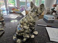 Front-right angle view of the cat figurine showing the sitting posture, tabby markings with grey and white coloration, glass eyes, and detailed paint finish.
