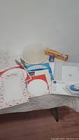Assorted baking supplies and accessories including decorative doilies, plastic cake carrier, dessert cups, scissors, plastic wrap, and cake decorating bags on a table.