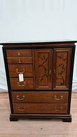 Front view of vintage Bassett chest showing birch wood drawers with brass hardware and black lacquered door panels featuring chinoiserie branch and floral design.