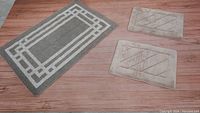 Three bath mats on wood floor: one large grey with white border pattern and two smaller beige mats with stitched line designs