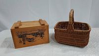 Photo showing a rectangular wooden box with an etched scene next to a woven basket with a tall handle.