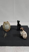 Four cat figurines displayed on dark grey cloth on white surface with white background. Items include a beige pottery cat, black ceramic cat, porcelain Siamese cat, and chrome ring holder cat.